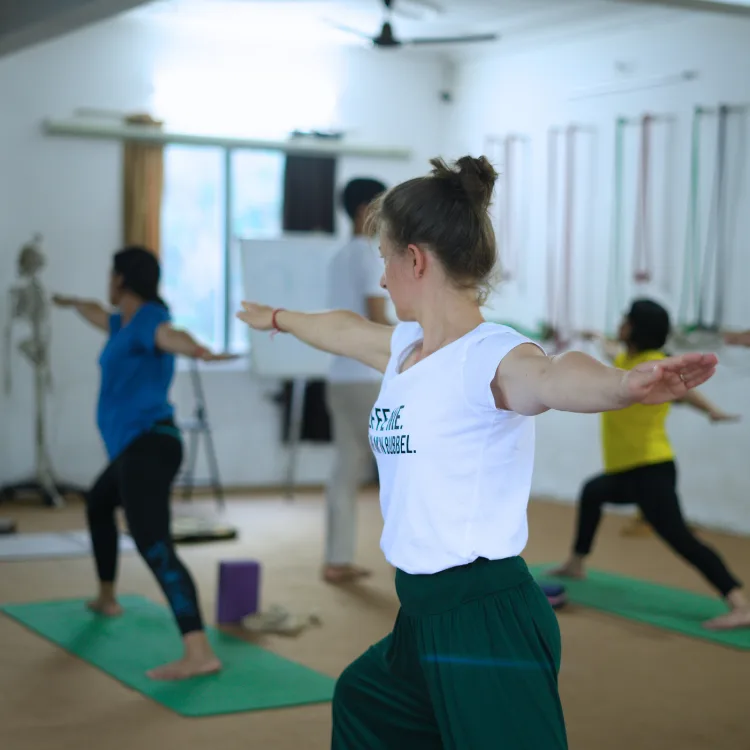 Students practicing Warrior II pose on mats during a 50 hour yoga teacher training in Rishikesh inside a yoga studio with props and alignment tools.