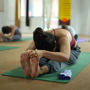 Female yoga student holding a seated forward bend pose (Paschimottanasana) on a green mat inside the shala during a 500 hour yoga teacher training in rishikesh practice class.