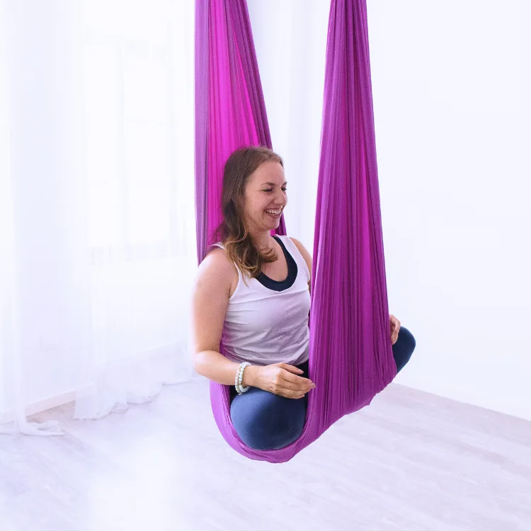 A smiling student practicing a seated pose in a purple hammock during an aerial yoga teacher training in Rishikesh in a bright studio.