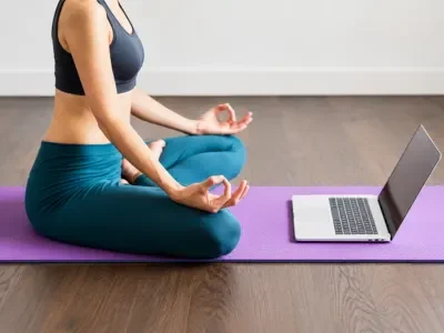 Woman practicing seated meditation on a yoga mat beside a laptop during a 200 Hour Online Yoga TTC